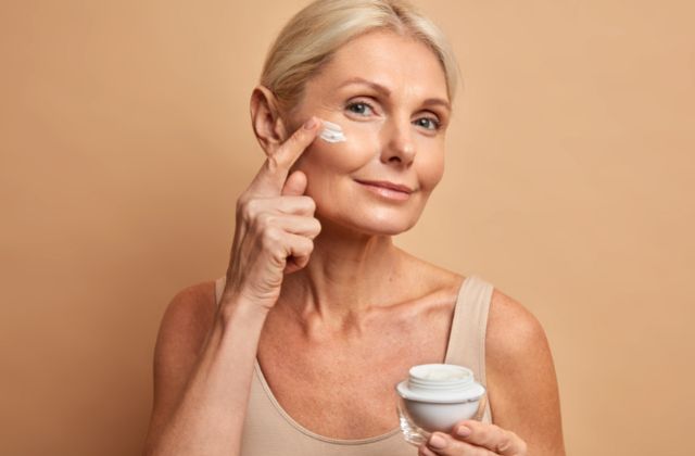 Mulher de meia idade loira de pele clara passa hidratante facial com a ponta dos dedos, segurando o produto na outra mão