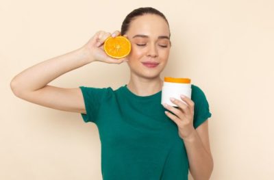 Mulher segurando uma laranja, representando a vitamina C, e um produtos para a pele